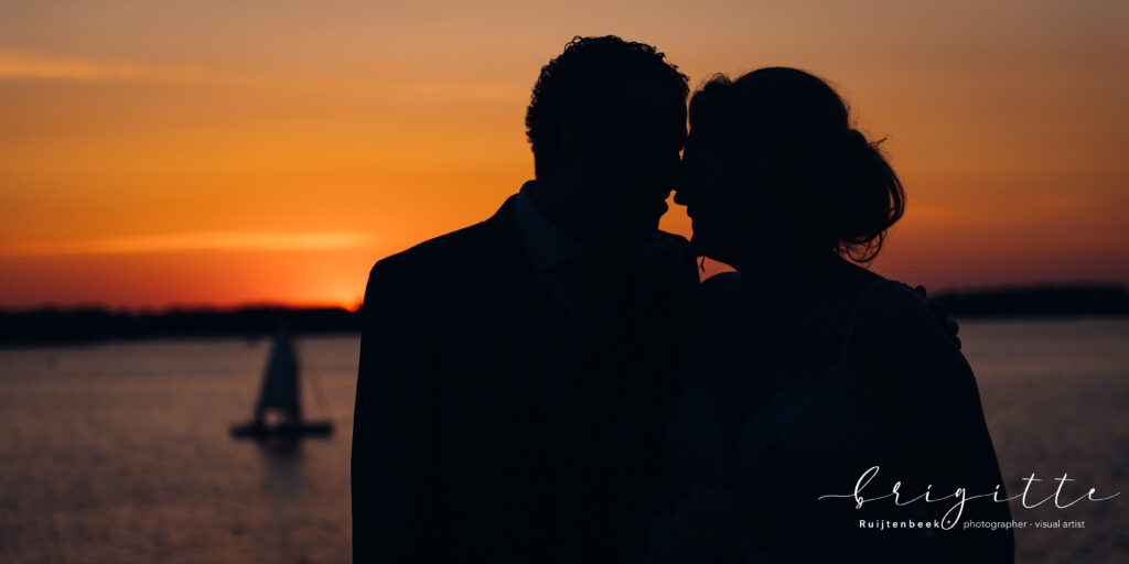 Sunset foto aan het water met bruidspaar als silhouet op de voorgrond, de lucht is oranje gekleurd