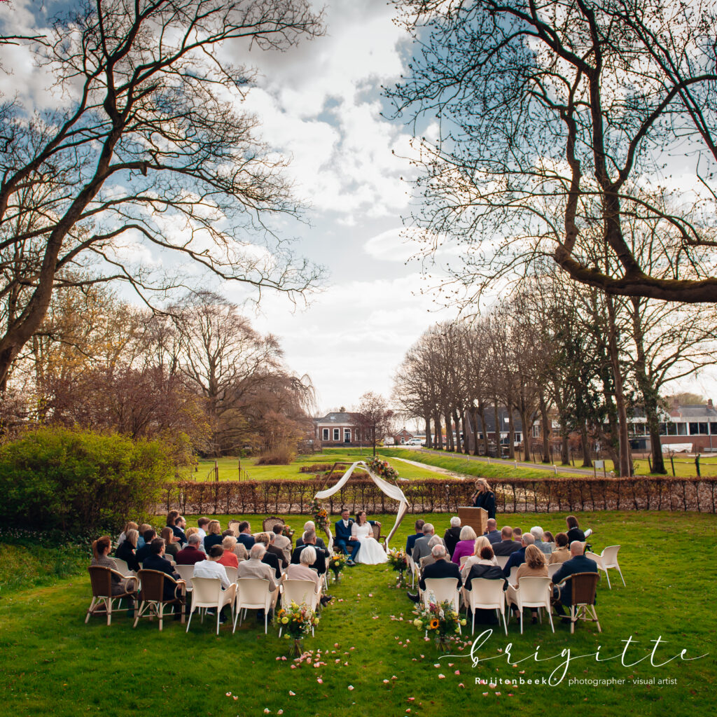 Overzichtsfoto van ceremonie buiten op het gras. De gasten zitten op versierde stoelen met hun gezicht naar het bruidspaar die op een bakje zit onder een met bloemen versierde boog