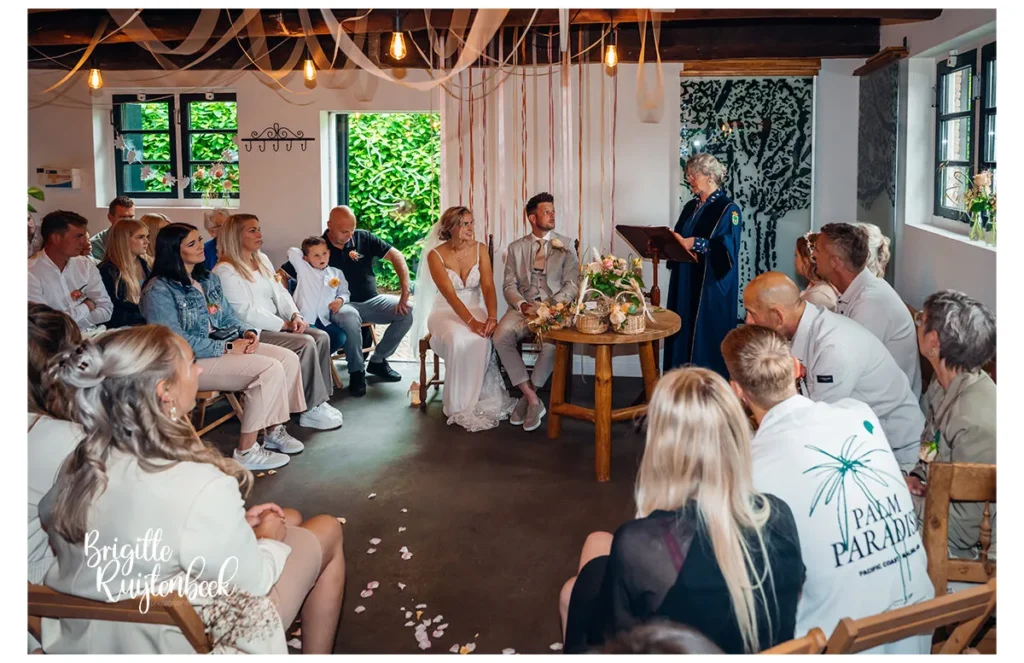 Bruidspaar zit tijdens de ceremonie binnen bij Slotplaats Bakkeveen, met familie en trouwambtenaar.