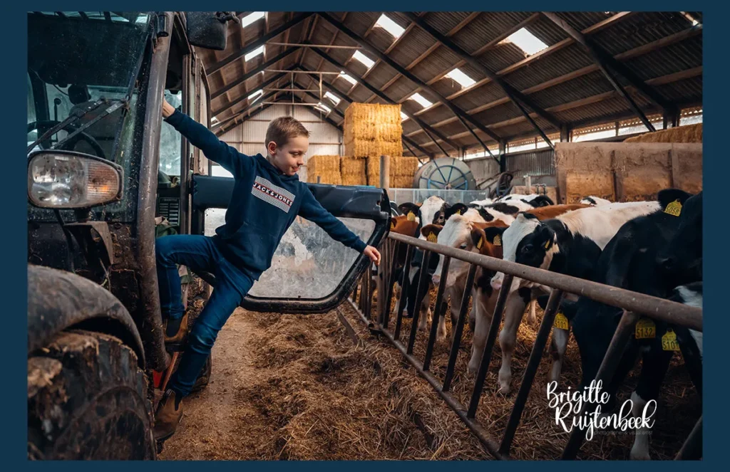 Jongen in boerenkleding stapt uit een trekker in de stal, met nieuwsgierige kalfjes op de achtergrond op een boerderij in Groningen.