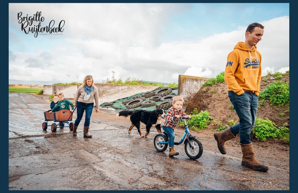 Gezin op een boerderij in Groningen: moeder trekt een bolderkar met twee kinderen terwijl de jongste voorop fietst, vader en hond lopen mee.