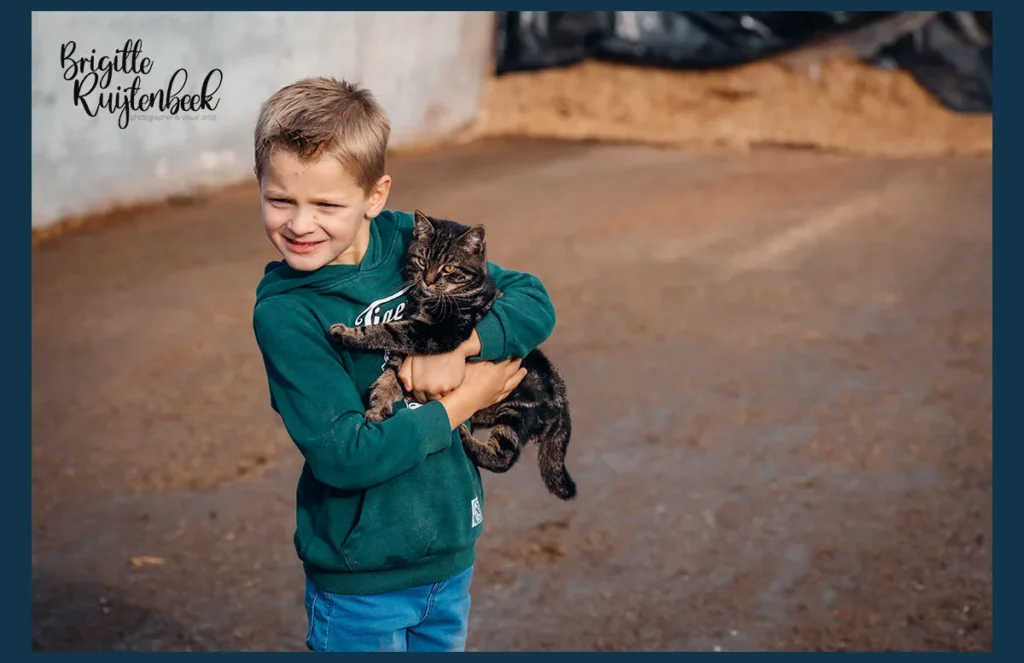 Jonge jongen in groene hoodie houdt een boerderijkat stevig vast tijdens een familiereportage op het erf van een boerderij in Groningen.