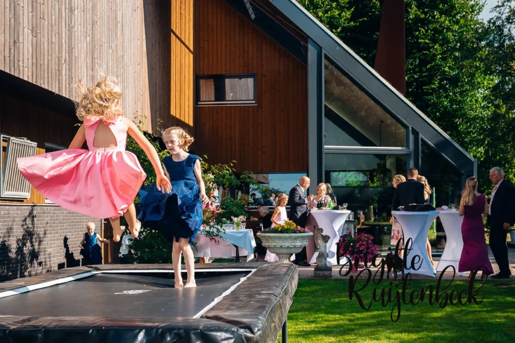 Twee jonge meisjes in feestjurken springen op een trampoline tijdens een bruiloftsreceptie in de tuin bij de boerderij in Kolham.