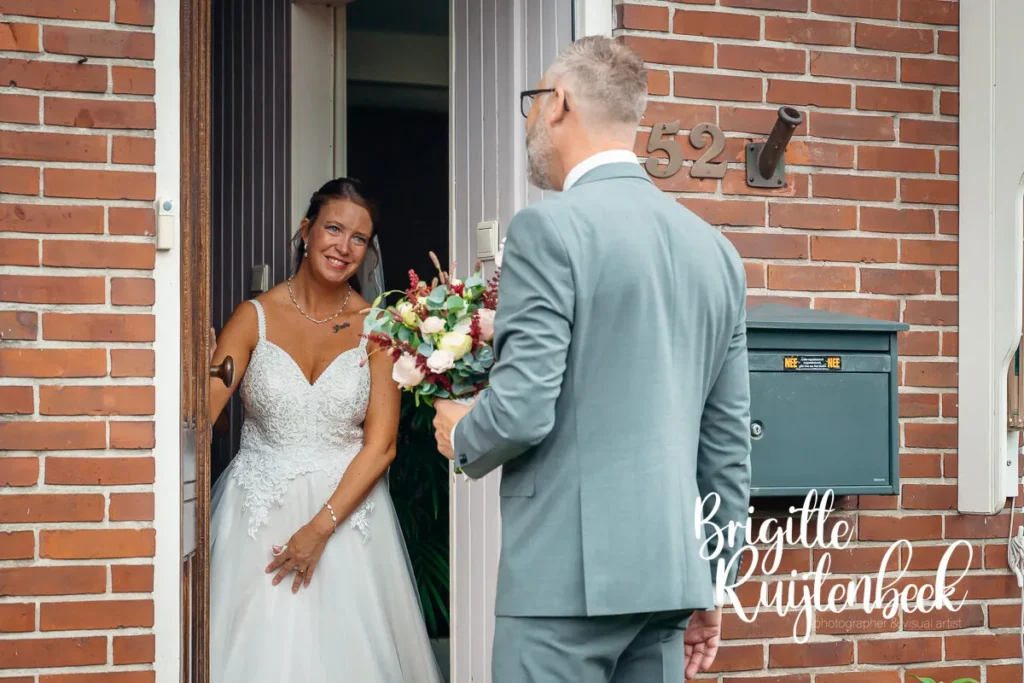 Bruid opent glimlachend de deur voor haar bruidegom met trouwboeket op hun trouwdag bij de Fraeylemaborg Slochteren.