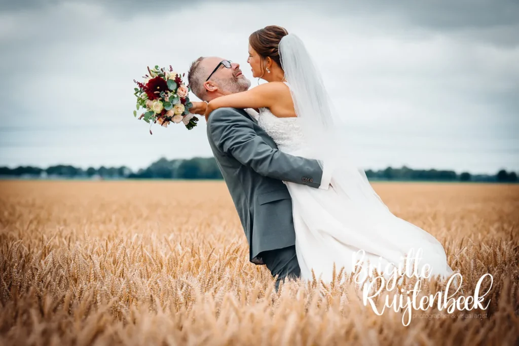 Bruidegom tilt bruid op in een gouden graanveld, lachend naar elkaar, op hun trouwdag bij de Fraeylemaborg.