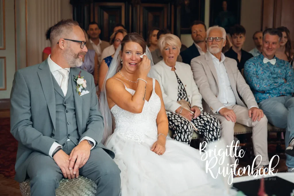 Bruid in witte trouwjurk pinkt ontroerd een traan weg tijdens de ceremonie bij de Fraeylemaborg in Slochteren, geflankeerd door de bruidegom en gasten.