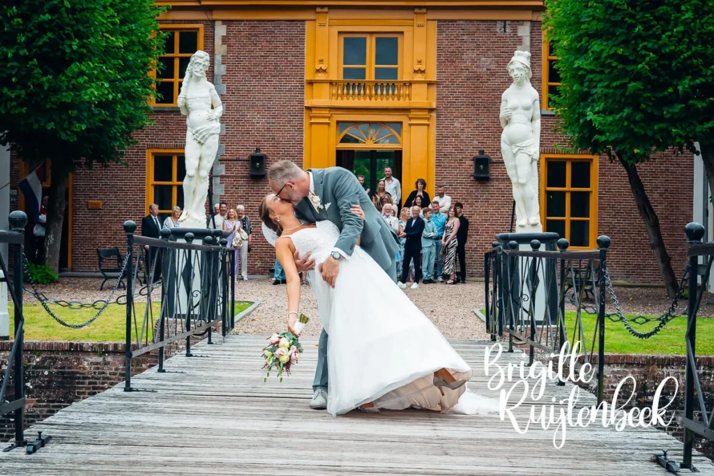 Bruid en bruidegom delen een romantische kus op de brug voor de Fraeylemaborg, omringd door juichende gasten.