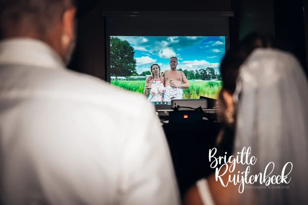 Bruidspaar kijkt lachend naar een humoristische videoboodschap van vrienden, geprojecteerd tijdens het feest bij Restaurant de Boerderij in Slochteren.