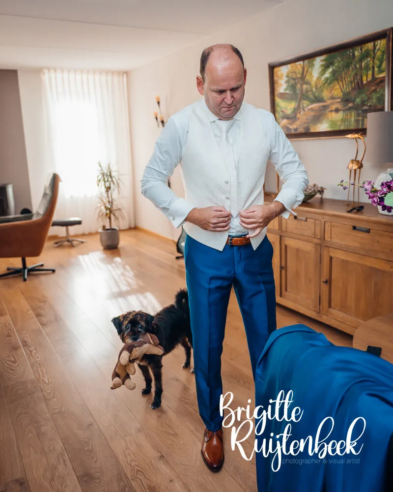 Bruidegom knoopt zijn gilet dicht terwijl zijn hond met knuffel in de bek toekijkt tijdens de voorbereidingen voor de bruiloft bij Trouwboerderij Mausel in Noordbroek.