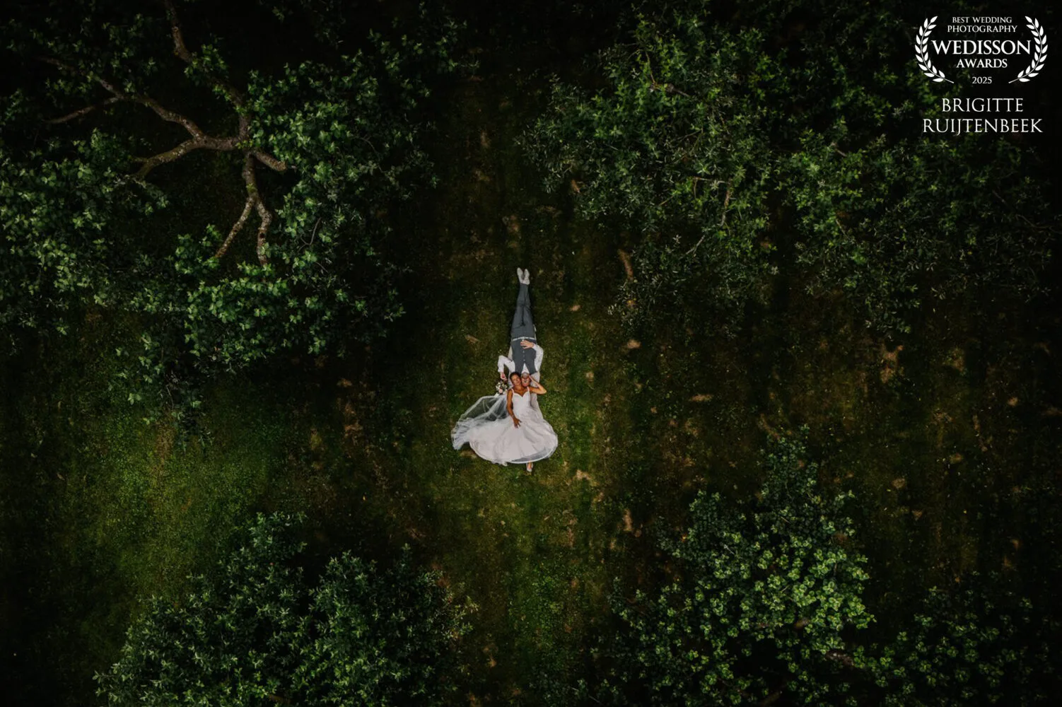 Adembenemende dronefoto van een bruidspaar dat samen op het gras ligt in een boomgaard, vastgelegd vanuit de lucht.