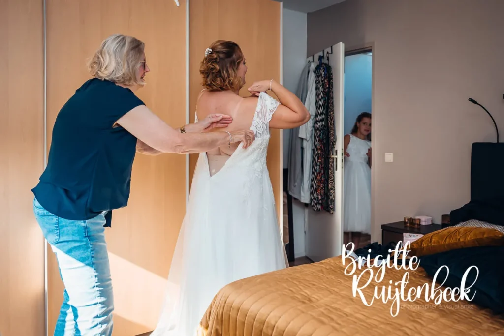 Moeder helpt de bruid bij het aantrekken van haar trouwjurk, terwijl een meisje nieuwsgierig meekijkt vanuit de deuropening, op de ochtend van de bruiloft bij Trouwboerderij Mausel in Noordbroek.
