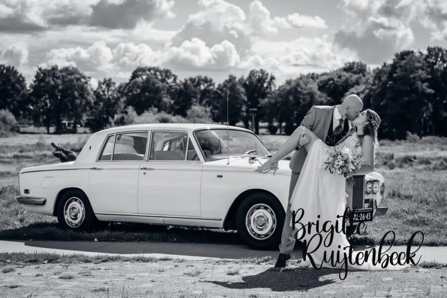 Bruidsfotografie De Wiemel Gasselte, bruid en bruidegom bij klassieke oldtimer in zwart-wit, kusmoment na de ceremonie.