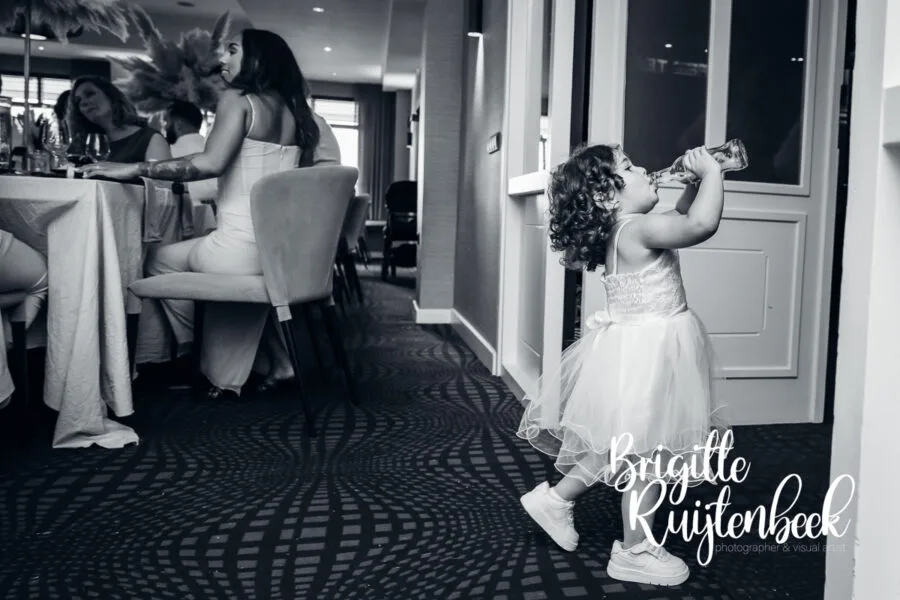 Jong kind in feestjurk drinkt uit een flesje terwijl gasten aan tafel zitten tijdens een bruiloft